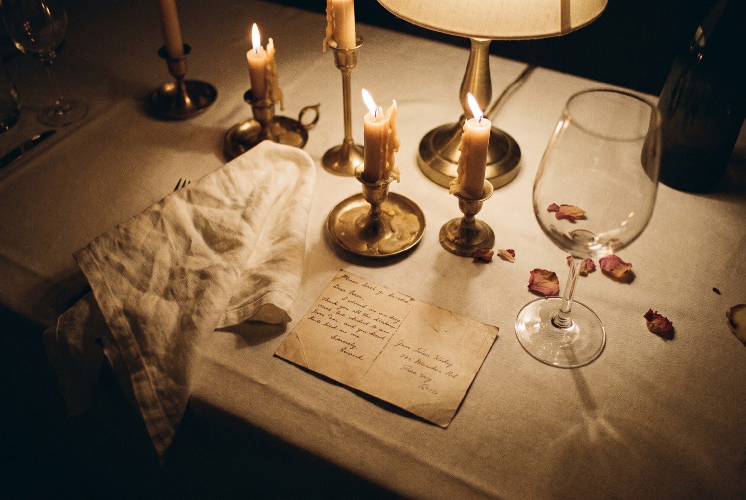 Een gedekte tafel laat op de avond: opgebrande kaarsen, een opgevouwen servet, een handgeschreven kaartje en een leeg glas.