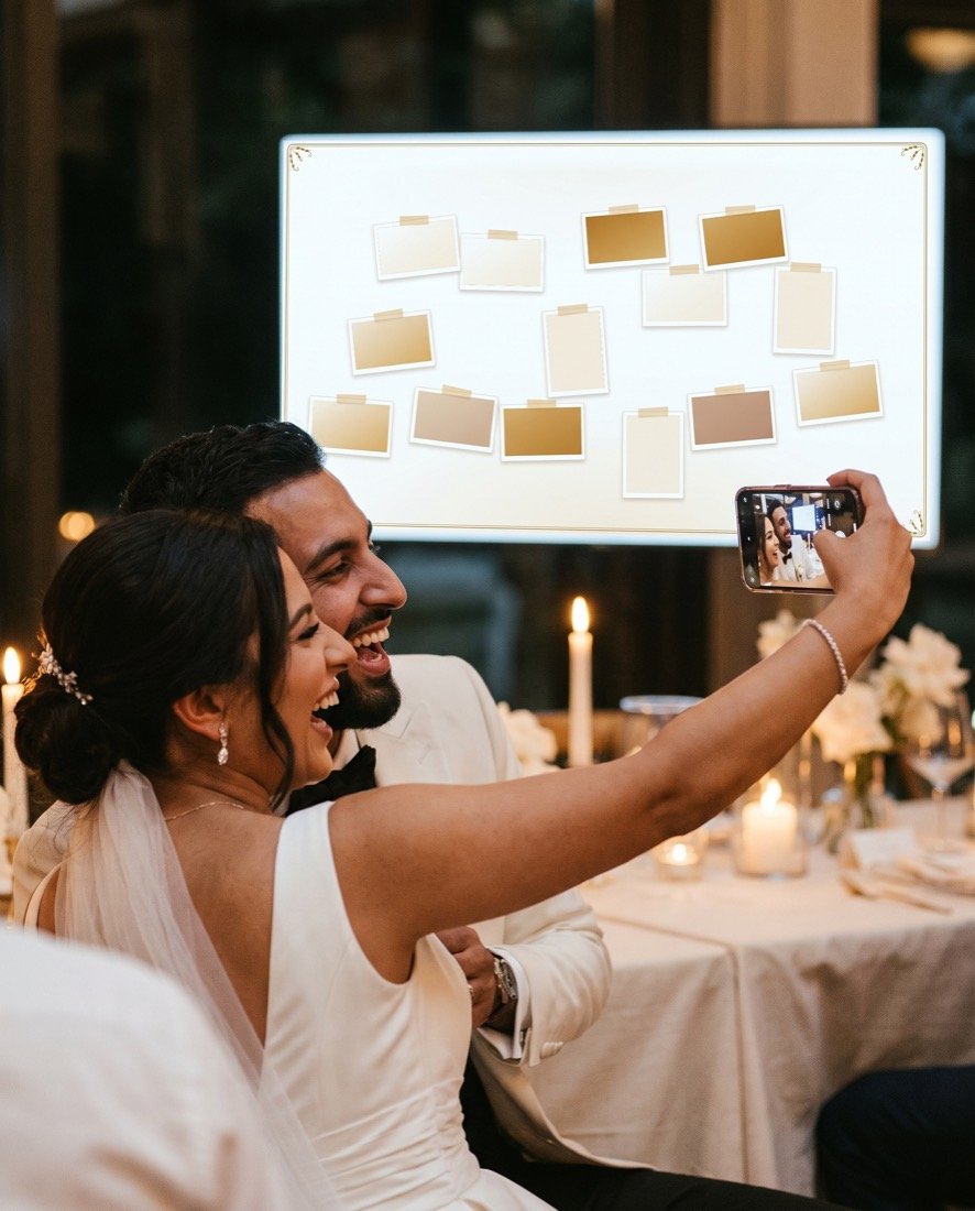 Een stel houdt een telefoon op armlengte om samen een selfie te maken aan een gedekte tafel, met de verlichte fotowand op de achtergrond.
